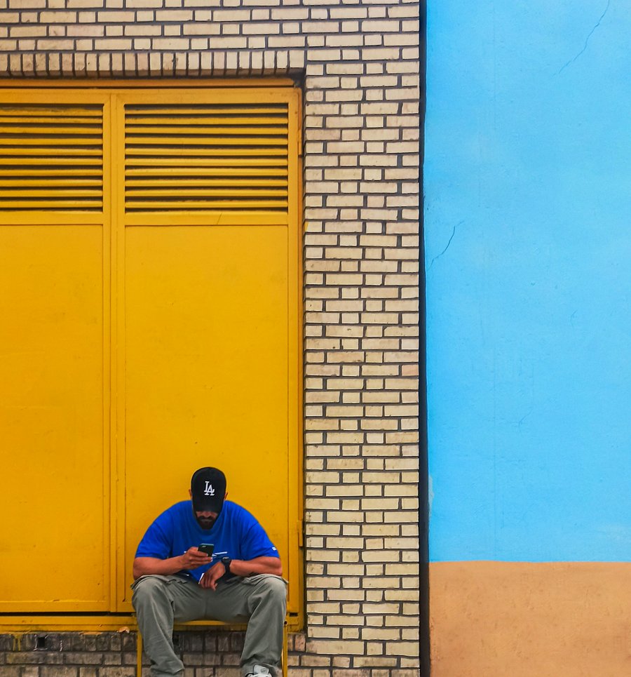 A man sitting by a bright yellow door on a blue wall, looking at his phone