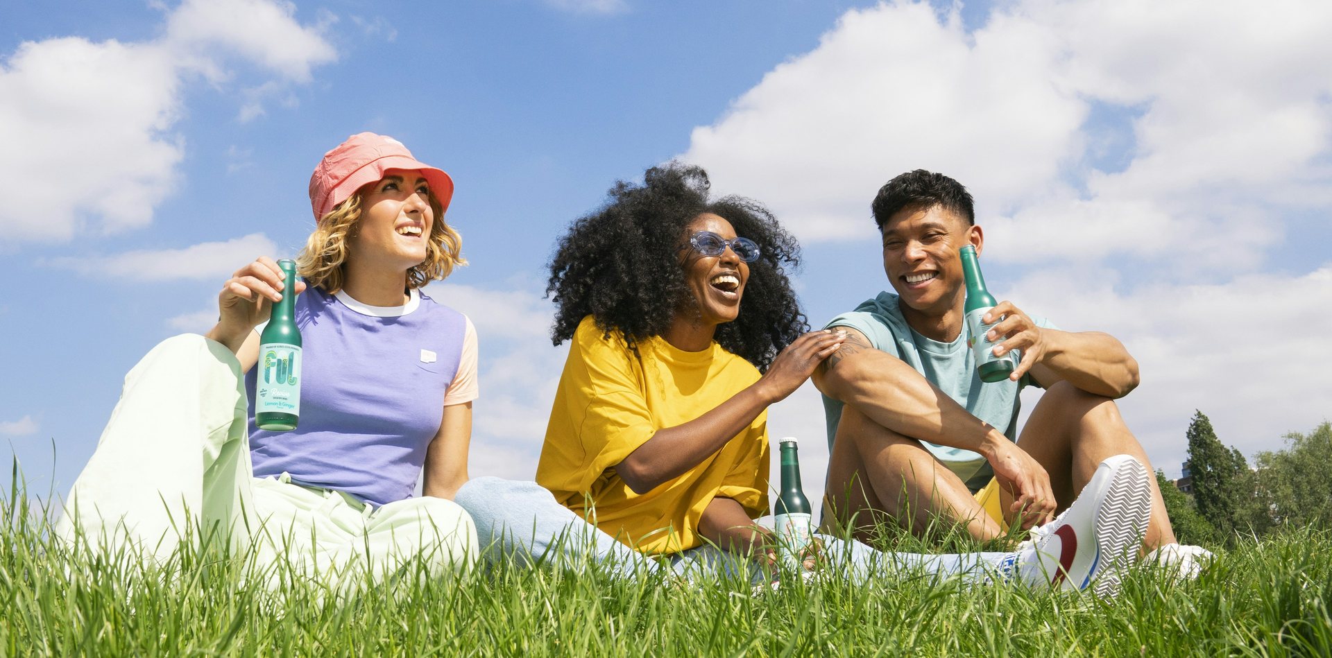Three friends laughing on the grass under a blue sky